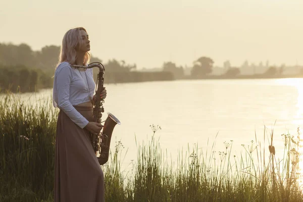 Silueta de una joven hermosa chica tocando el saxofón al amanecer junto al río, una mujer en un vestido largo en la naturaleza al amanecer relajante, música conceptual — Foto de Stock