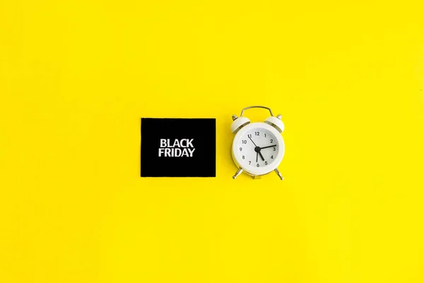 white alarm clock and an inscription on a black square - black Friday on a yellow background. The topic of sales. Flat lay.
