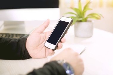 Adam masaüstünde çalışırken bir eliyle bir telefon tutuyor.