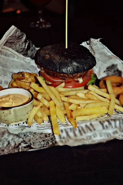 Burger of black bun with tomato, juicy beef cutlet, serving on newspaper with cheese sauce and French fries