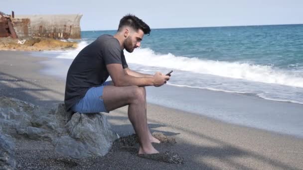 Человек занят своим телефоном, сидящим на пляжных валунах — стоковое видео