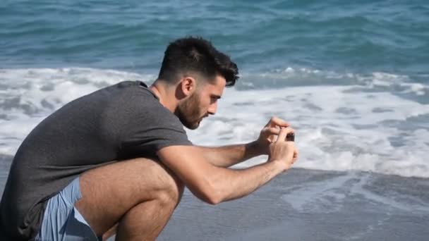Молодой человек использует мобильный телефон, чтобы сфотографироваться на пляже — стоковое видео