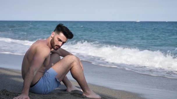 Молодий чоловік сидить на пляжі, граючи з піском — стокове відео