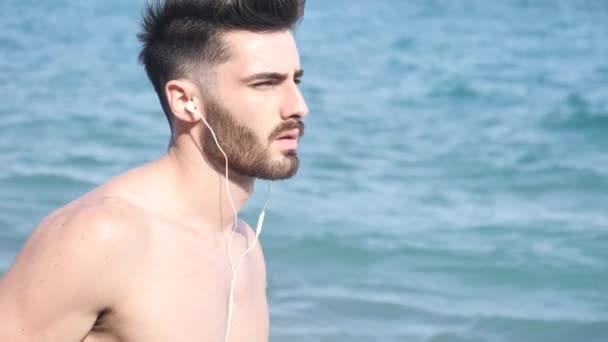 Sportlicher junger Mann am Strand und hört Musik — Stockvideo