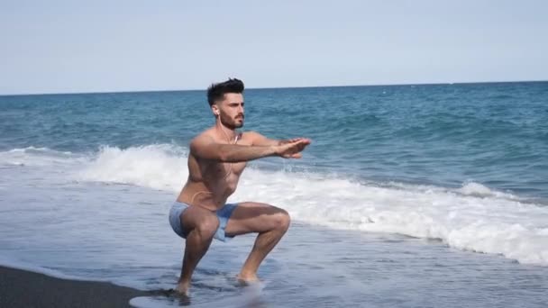 Junger Mann turnt, macht Kniebeugen am Strand — Stockvideo