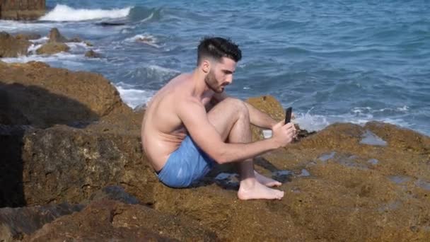 Hombre joven en la playa usando el teléfono celular para filmar el mar — Vídeo de stock