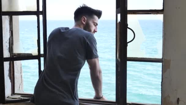 Jonge man leunt tegen venster met zee achter — Stockvideo
