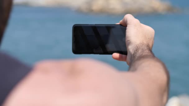 Νεαρός άνδρας που χρησιμοποιούν κινητό τηλέφωνο για να τραβήξετε φωτογραφία στην παραλία — Αρχείο Βίντεο