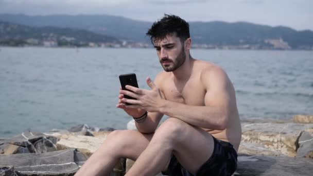 Jonge man op strand met behulp van de mobiele telefoon naar de film van de zee — Stockvideo