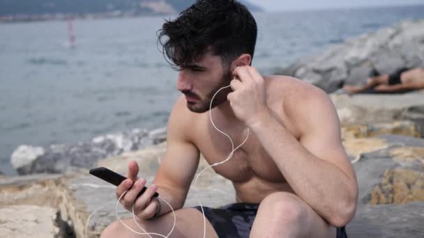 イヤホンで音楽を聴くビーチで若い男 — ストック動画