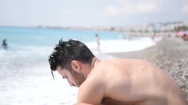 Молодой человек сидит на пляже — стоковое видео
