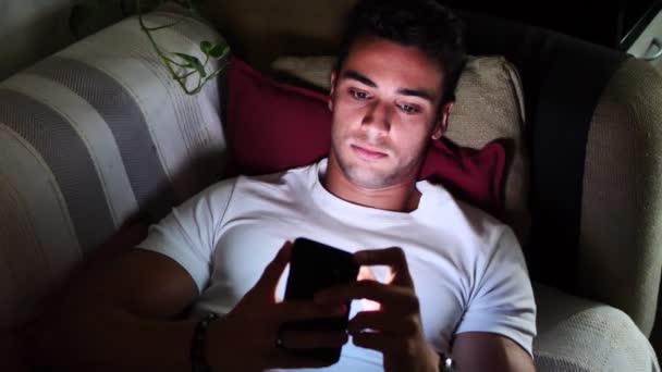 Joven en Sofá Lit por la luz de la pantalla del teléfono celular — Vídeos de Stock