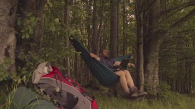 Adam gün batımında hamakta dizüstü bilgisayarla dağlarda çalışıyor.