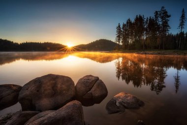 Kozalaklı ağaçlarla çevrili bir dağ gölü üzerinde güneş ışınlarıyla güzel bir gün doğumu manzarası. Bulgaristan 'ın Rodopi Dağları' ndaki Shiroka Polyana barajı.