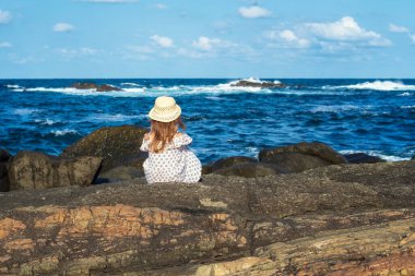 Küçük güzel bir kızın arkası kayalık bir sahil şeridinde güneşli ve sıcak bir günün tadını çıkarıyor.