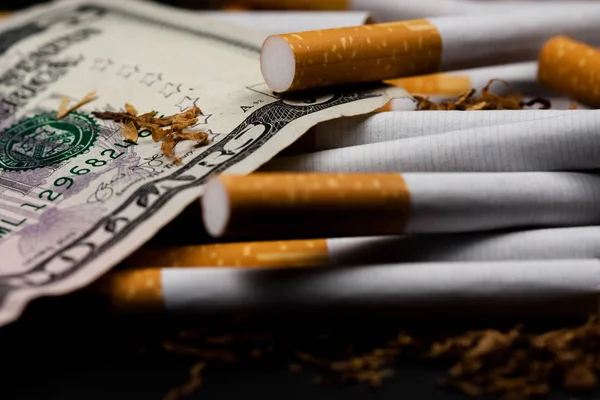 Cigarros separados com algum tabaco desfiado espalhado, com centavos ao redor e alguns dólares, isolados em fundo preto. O tabaco pode causar doenças . — Fotografia de Stock
