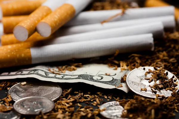Сигарети розділені з розкиданим тютюном, з копійки навколо і деякими доларами, ізольовані на чорному тлі. Тютюн може спричинити захворювання . — стокове фото
