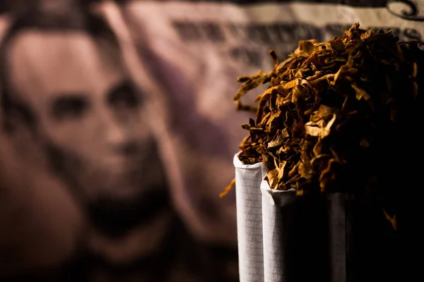 Numerous isolated tobacco cigarettes with shredded tobacco on top of them on a background with a five dollar bill. Tobacco can cause diseases.