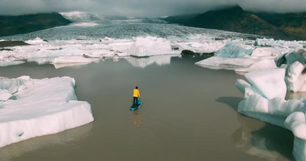 アイスランドで巨大氷山と氷河のラグーンでパドルボード上を立ってをパドリング明るい黄色のレインコートを着た男の空撮 — ストック動画