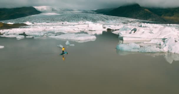 アイスランドで巨大氷山と氷河のラグーンでパドルボード上を立ってをパドリング明るい黄色のレインコートを着た男の空撮 — ストック動画