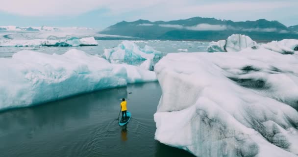 アイスランドで巨大氷山と氷河のラグーンでパドルボード上を立ってをパドリング明るい黄色のレインコートを着た男の空撮 — ストック動画