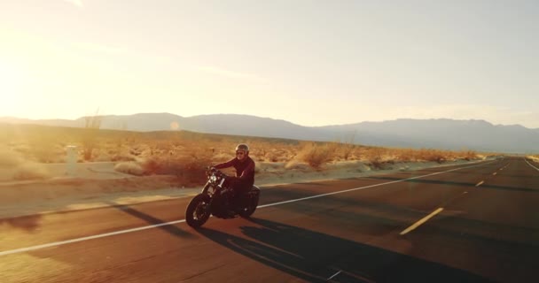 Mladý muž na koni vintage černý motocykl po Pouštní silnici při západu slunce