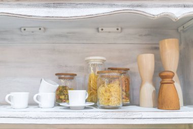 Beyaz ahşap mutfak dolabında beyaz seramik kahve fincanı ve şişedeki çeşitli gıda malzemeleri yiyecek ve içecek konseptidir.