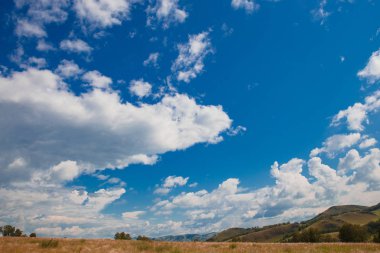 Beyaz bulutlar, ağaçlar, tarlalar ve yeşil çimenler ile çayırlar ile mavi gökyüzü, dağlara karşı. Doğanın bileşimi. Kırsal yaz manzarası.