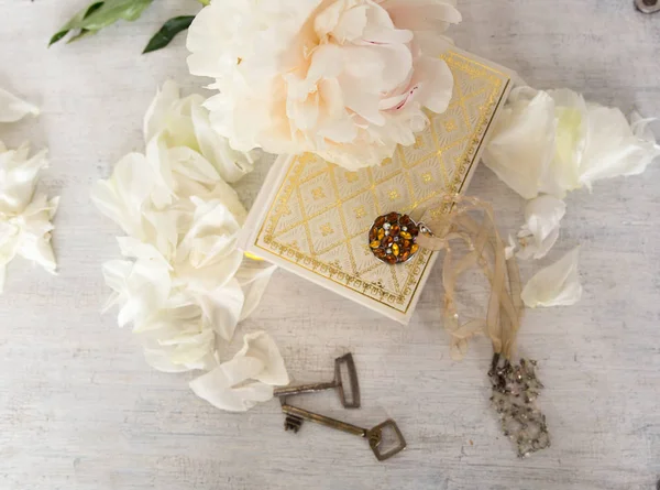 一幅美丽的白牡丹躺在一本有金色页面的书上。附近是精致的花瓣,复古的钥匙和装饰在脖子上。浅白色和米色复古背景. — 图库照片