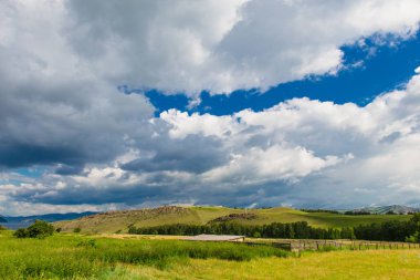Beyaz bulutlar, ağaçlar, tarlalar ve yeşil çimenler ile çayırlar ile mavi gökyüzü, dağlara karşı. Doğanın bileşimi. Kırsal yaz manzarası.