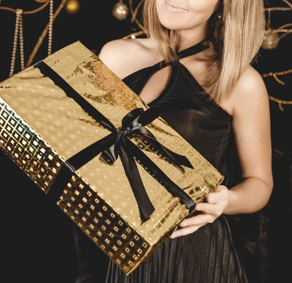 Siyah bir gece elbisesi içinde güzel bir gülümseme ile bir kız, siyah bir kurdele ile altın bir paket içinde bir hediye tutan. Büyük hediye kutusu. Siyah ve koyu arka plan.