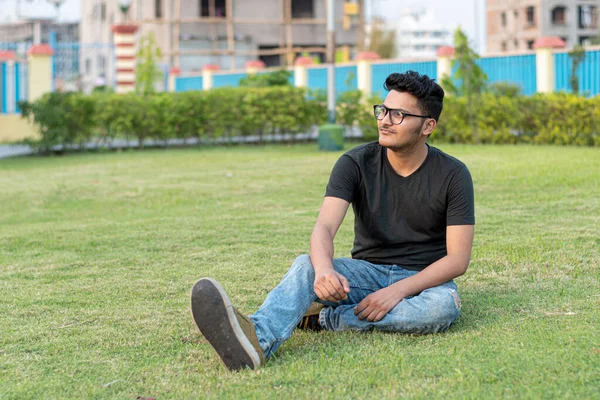 Fashion portrait of Indian Guy wearing a black t shirt, sits on the ground and poses against the background of nature. Indian lifestyle and fashion