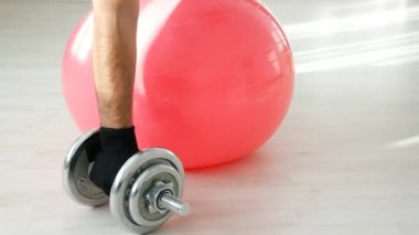 Mavi spor ayakkabılı ve eldivenli sporcuların elleri kırmızı topa yaslanmış ve dambılları kaldırıyor.
