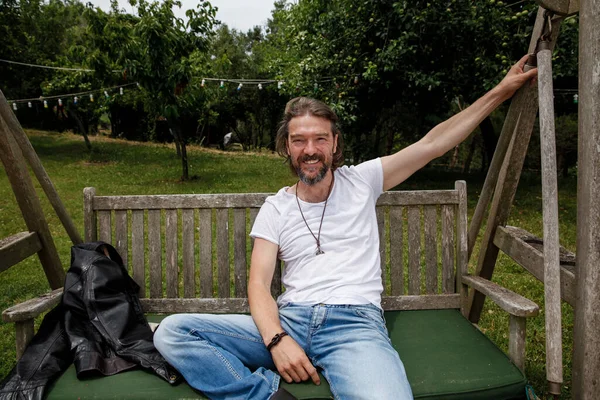Portrait of a handsome man sitting on a wooden garden swing seat in a natural parkland and smiling. Bearded man wearing white T-shirt and denim jeans.