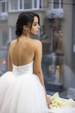 Parlak straplez balo elbisesi içinde zarif bir şekilde pencerenin yanında çiçeklerle poz veren zarif bir gelin.