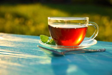 Mavi ahşap masa üzerinde nane yaprağı ve bir kaşık ile çay şeffaf bir cam fincan, bulanık yeşil doğal arka plan, güneşli bir gün