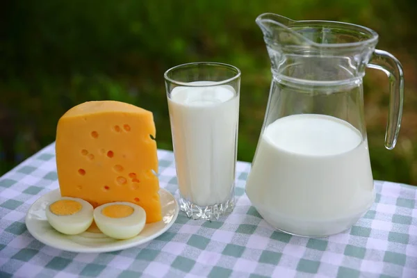 ミルクジャグ ミルクのグラス および緑と白のチェックテーブルクロスにカット卵とチーズの一部の朝食の組成物 ぼやけた緑の自然な背景 — ストック写真