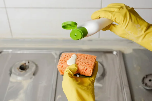 Two hands applying liquid soap from cleanser bottle to orange sponge. Extruding detergent onto orange sponge on background of gas stove.