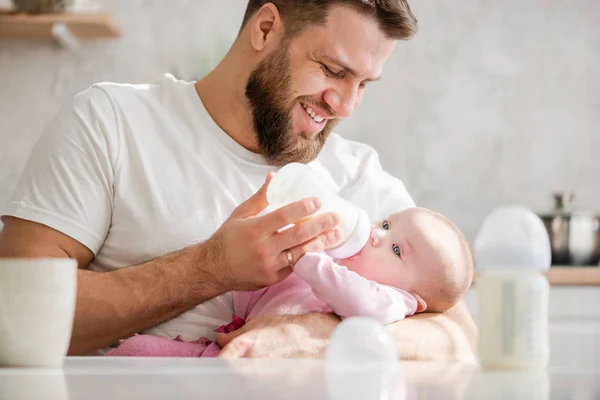 Feeding newborn with formula in a bottle in kitchen.