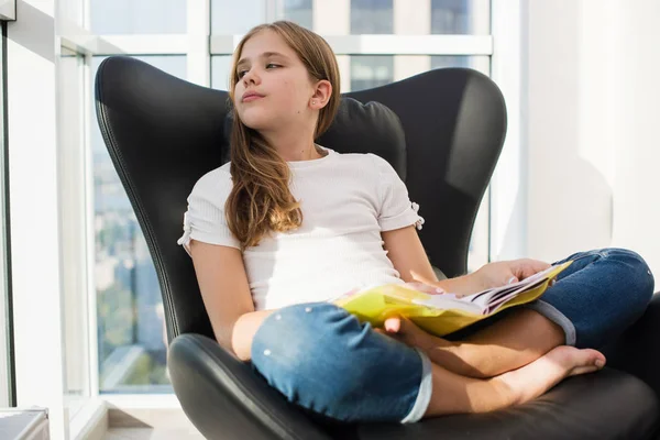 Soñando niña de la escuela sentarse en el sillón de casa con libro — Foto de Stock