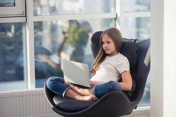 Linda chica de la escuela sentarse en un sillón acogedor con ordenador portátil — Foto de Stock