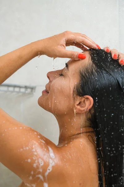 若い美しいブルネットの女性の側の肖像画は 水の滴と泡で首 顔と髪を洗うために蛇口の下に頭を上げる シャワーを浴びます 白いタイルバスルーム — ストック写真