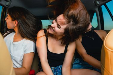 Arabadaki üç genç bayan kuaförde arka koltukta oturuyorlar, müzik dinliyorlar, eğleniyorlar, dans ediyorlar, el sallıyorlar ve kaygısız davranıyorlar.