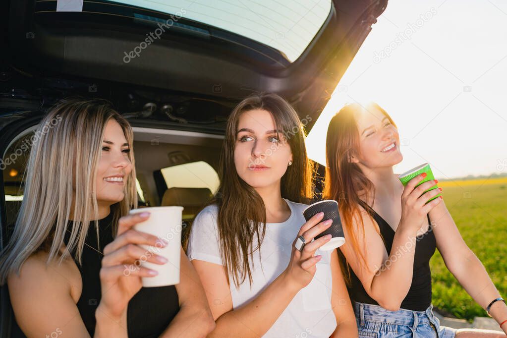 Three girls have coffee break at car trunk. Happy road trip smiling female travelers, relax at vehicle back with coffee cups take away at sunlight.