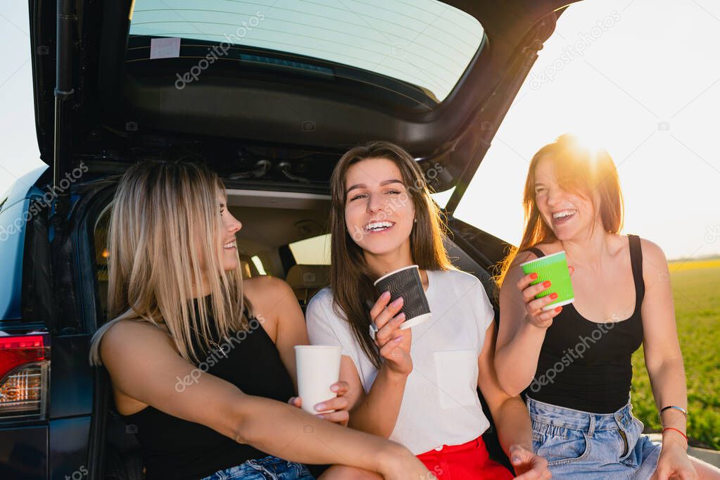 Happy road trip of three smiling women, relax at vehicle back with coffee cups take away at sunlight.
