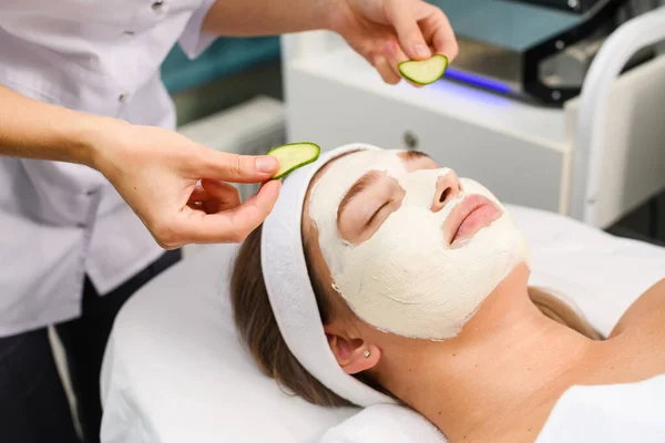 Beautician apply natural white moisturizing face mask and refresh eyes area with cucumber, skincare concept, professional treatment for lying and relaxing female patient