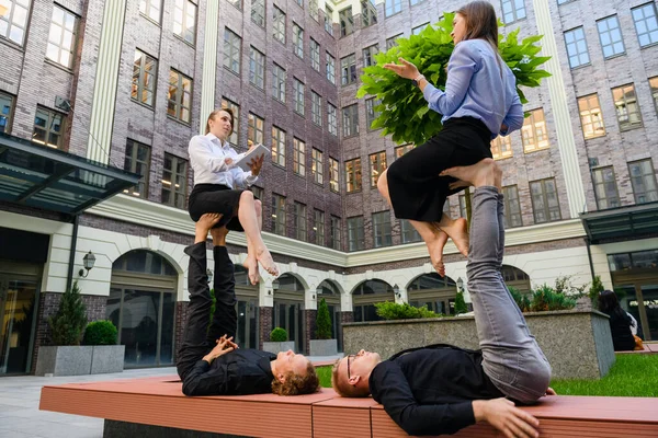 Acroyoga concept with four people, two base men lay on bench at hold at feet two flyer girls during work talking, take note, interview in office clothes