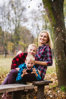 Sarışın aile sonbahar parkında ahşap bankta oynuyor, anne iki ergen erkek ikizi kucaklıyor ve gülümsüyor, açık havada aktif bir hafta sonu.