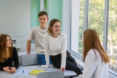 Parlak sınıfta gülümseyen ve sohbet eden bir grup genç lise öğrencisi, pencereden gelen doğal ışıkla rahatlatılmış bir atmosfer..