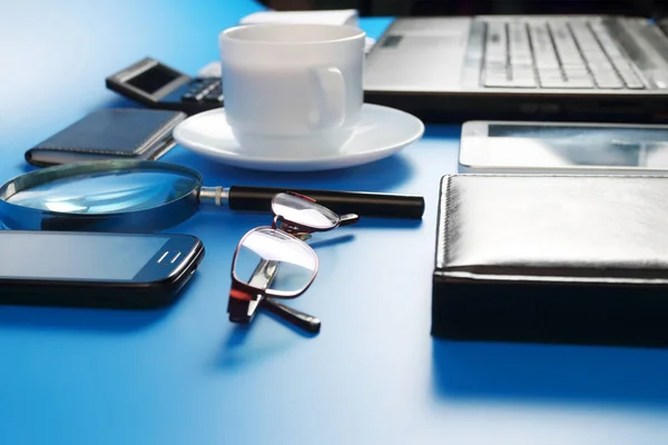 Tablet PC, laptop, mobile phone, glasses and cup with black coffee on saucer on blue background